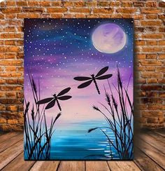 A vertical canvas wall art print featuring a night sky with a large moon, stars, two dragonflies, and silhouetted reeds over blue water, displayed against a brick wall and wood floor.
