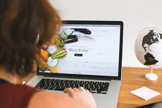 a woman looks at a lap top displaying a Black Friday promo screen on a wooden desk with a black and white globe and blank notepad