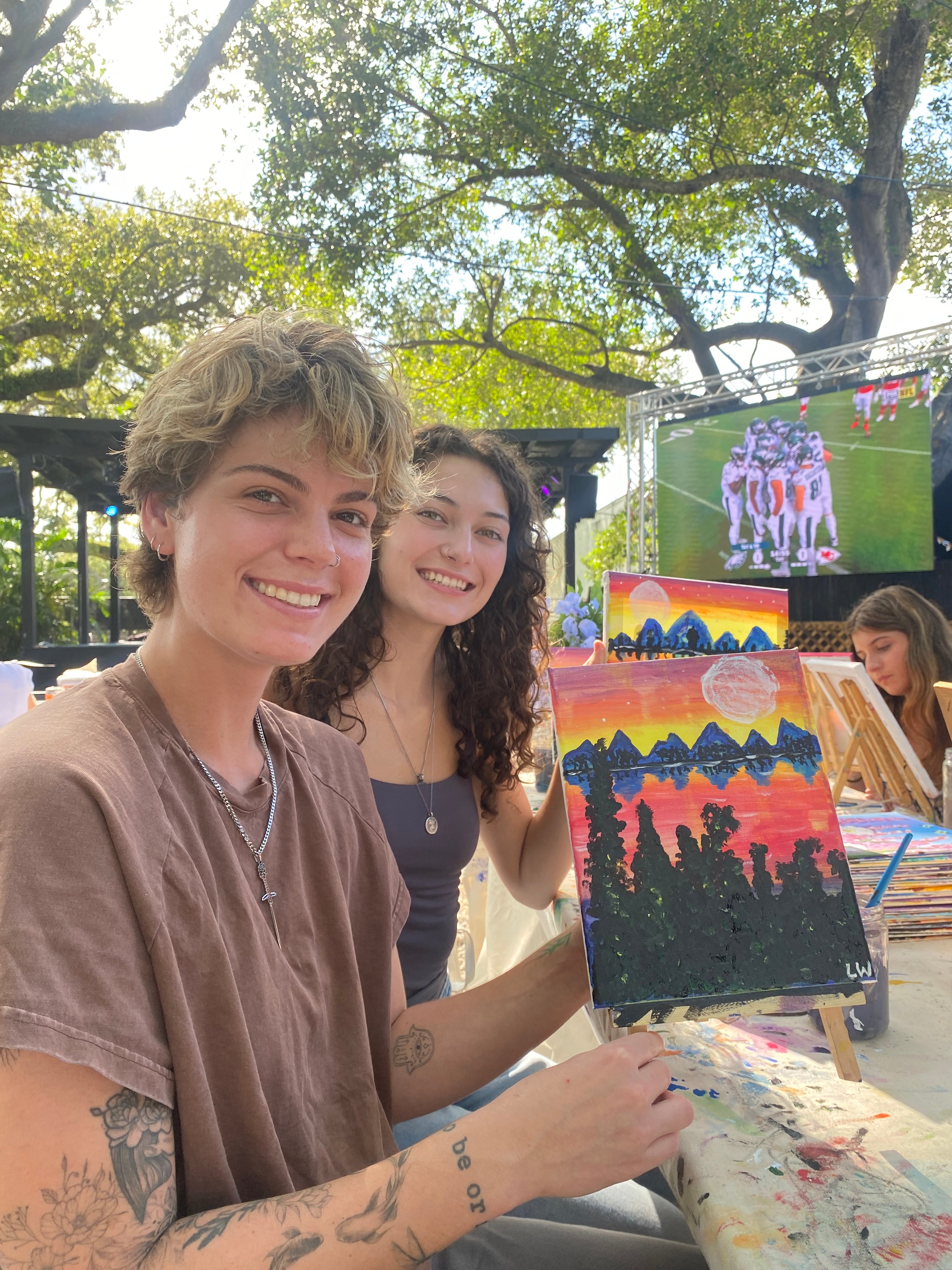 Two people with art supplies outdoors, surrounded by trees.